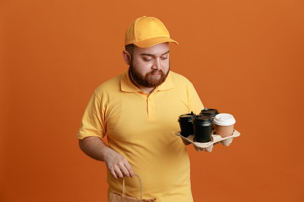 Delivery man employee in yellow cap blank tshirt uniform holding coffee cups and paper bag looking at cups with serious face standing over orange background