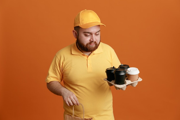 Сотрудник службы доставки в желтой кепке с пустой футболкой держит кофейные чашки и бумажный пакет, глядя на чашки с серьезным лицом, стоящим на оранжевом фоне