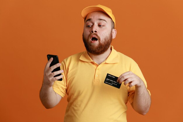 オレンジ色の背景の上に立って驚いている携帯電話を見てクレジットカードを保持している黄色のキャップの空白のTシャツの制服を着た配達人の従業員