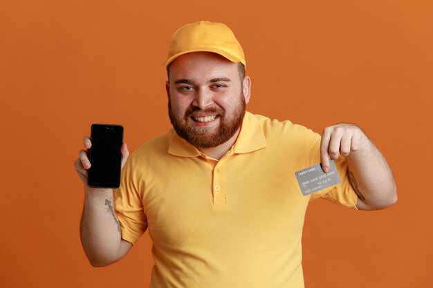 オレンジ色の背景の上に立って幸せでポジティブな笑顔のカメラを見てスマートフォンを示すクレジットカードを保持している黄色のキャップの空白のTシャツの制服を着た配達人の従業員