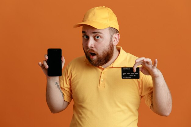 Delivery man employee in yellow cap blank tshirt uniform holding credit card showing smartphone looking at camera surprised standing over orange background