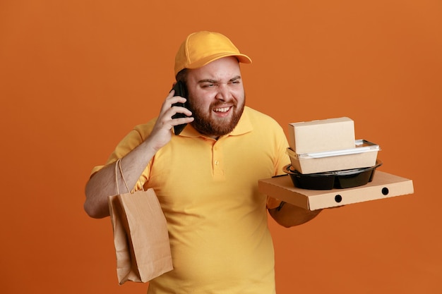 노란색 모자 빈 티셔츠를 입은 배달원 직원이 음식 용기와 피자 상자를 들고 휴대전화로 통화하는 종이 봉지가 오렌지색 배경 위에 서 있는 것을 짜증나게 한다