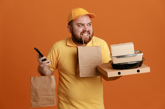 Сотрудник службы доставки в желтой кепке с пустой футболкой держит контейнер с едой и коробку для пиццы с бумажным пакетом в зубах, выглядя раздраженным и злым, стоя на оранжевом фоне