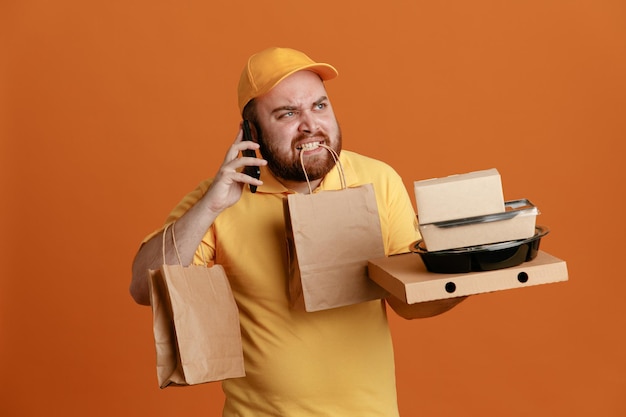 オレンジ色の背景の上に立っているイライラした表情で携帯電話で話している歯の紙袋と食品容器とピザボックスを保持している黄色のキャップの空白のTシャツの制服を着た配達人の従業員