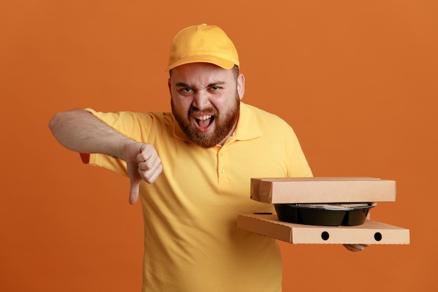 Сотрудник службы доставки в желтой кепке с пустой футболкой держит контейнер с едой и коробки для пиццы, глядя в камеру с сердитым лицом, показывая большой палец вниз, стоя на оранжевом фоне