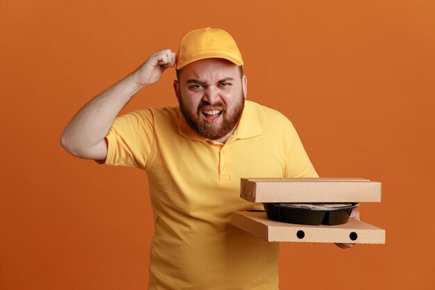 オレンジ色の背景の上に立っているイライラした表情で怒った顔でカメラを見て食品容器とピザの箱を保持している黄色のキャップの空白のTシャツの制服を着た配達人の従業員