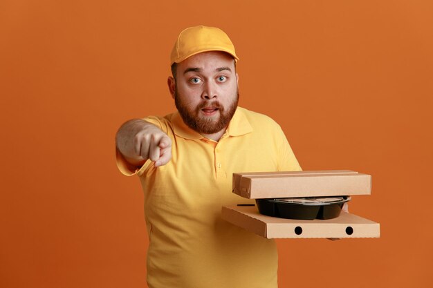 Delivery man employee in yellow cap blank tshirt uniform holding food container and pizza boxes pointing with index finger at camera being surprised standing over orange background