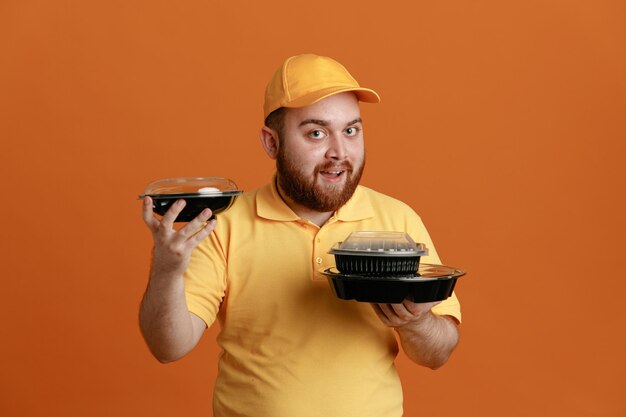 Delivery man employee in yellow cap blank tshirt uniform holding food containers looking at camera happy and positive smiling cheerfully standing over orange background