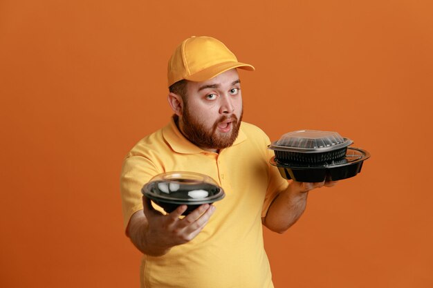 Delivery man employee in yellow cap blank tshirt uniform holding food containers looking at camera with confident expression standing over orange background