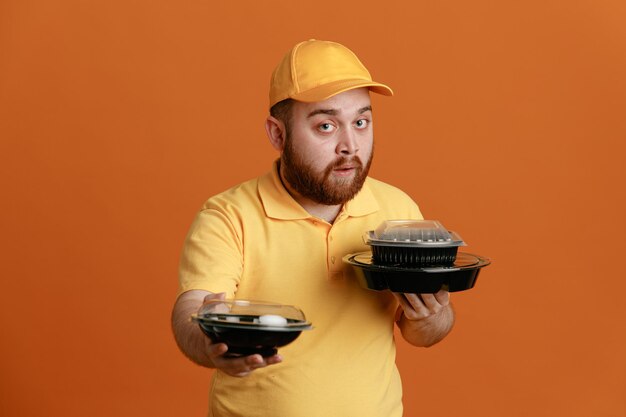 Delivery man employee in yellow cap blank tshirt uniform holding food containers looking at camera with confident expression standing over orange background