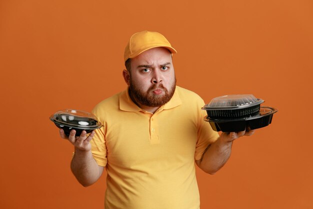 Delivery man employee in yellow cap blank tshirt uniform holding food containers looking at camera with frowning face standing over orange background