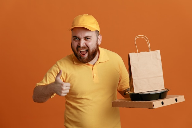 Сотрудник службы доставки в желтой кепке с пустой футболкой держит контейнеры с едой и коробку для пиццы с бумажным пакетом, смотрит в камеру счастливым и взволнованным, показывая большой палец вверх, стоя на оранжевом фоне