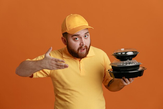 Delivery man employee in yellow cap blank tshirt uniform holding food containers presenting with hand looking confident standing over orange background