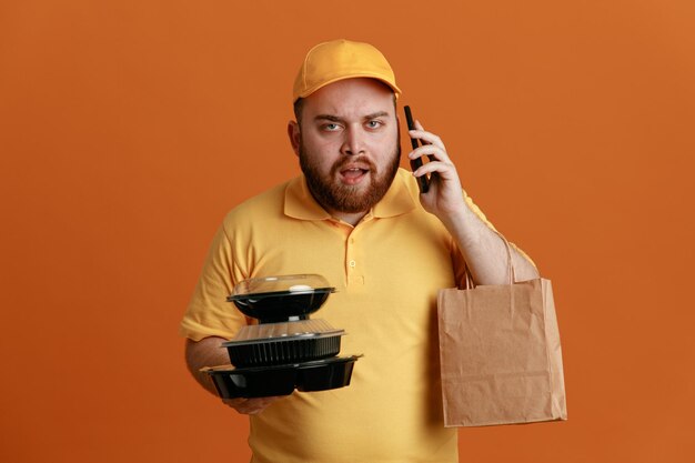 Delivery man employee in yellow cap blank tshirt uniform holding food containers with paper bag talking on mobile phone looking confused and displeased standing over orange background