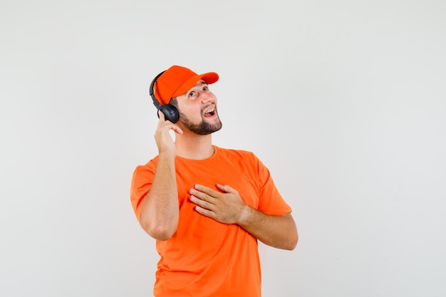 Доставщик, наслаждаясь музыкой с наушниками в оранжевой футболке, кепке и радостно глядя, вид спереди.
