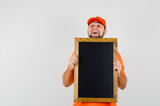 Доставка человек держит пустую доску в оранжевой футболке, кепке и выглядит веселым. передний план.