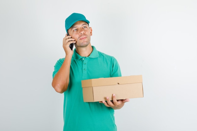 Delivery man holding box and talking on phone in green t-shirt with cap