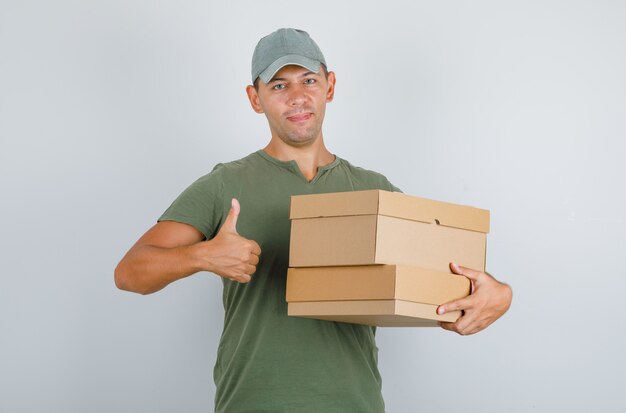 Delivery man holding boxes and showing thumb up in green t-shirt, cap. front view.