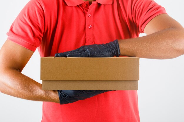 Delivery man holding cardboard box in red uniform, medical gloves , front view.