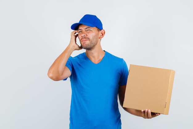 Delivery man holding cardboard box with fingers on face in blue t-shirt