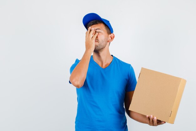 Delivery man holding cardboard box with hand on face in blue t-shirt