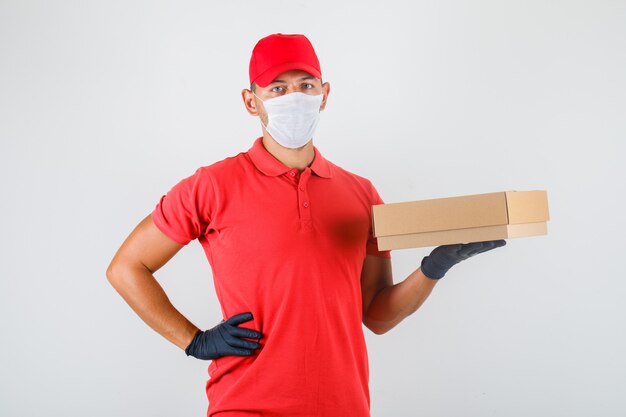 Delivery man holding cardboard box with hand on waist in red uniform, medical mask, gloves front view.