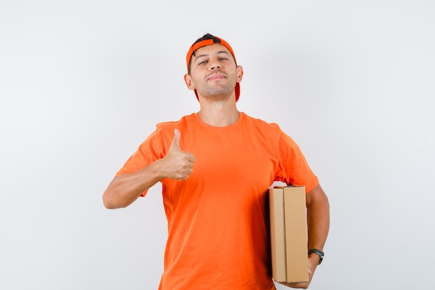 Delivery man holding cardboard box with thumb up in orange t-shirt and cap and looking confident