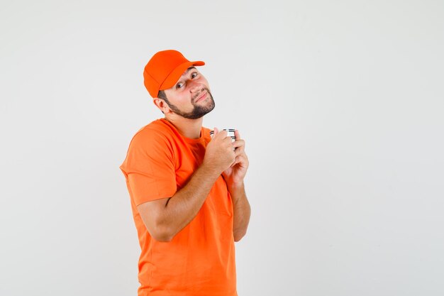 Delivery man holding cup of drink in orange t-shirt, cap and looking cute. front view.