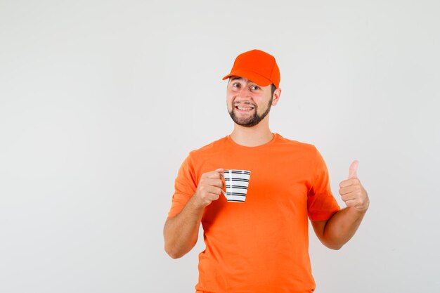 Доставщик еды, держащий чашку напитка с большим пальцем руки вверх в оранжевой футболке, кепке и смотрящий жизнерадостный, вид спереди.