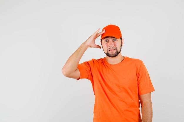 Delivery man holding fingers on head in orange t-shirt, cap and looking distressed. front view.