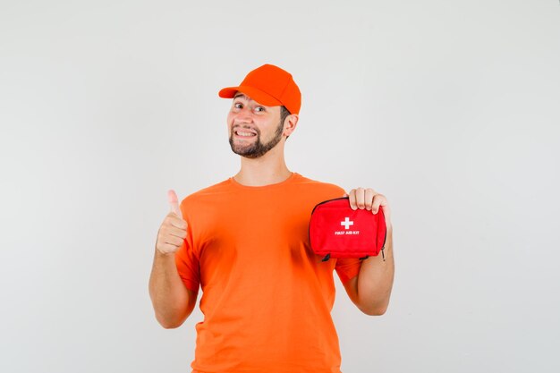 Fattorino che tiene il kit di pronto soccorso con il pollice in su in maglietta arancione, berretto e sembra allegro. vista frontale.