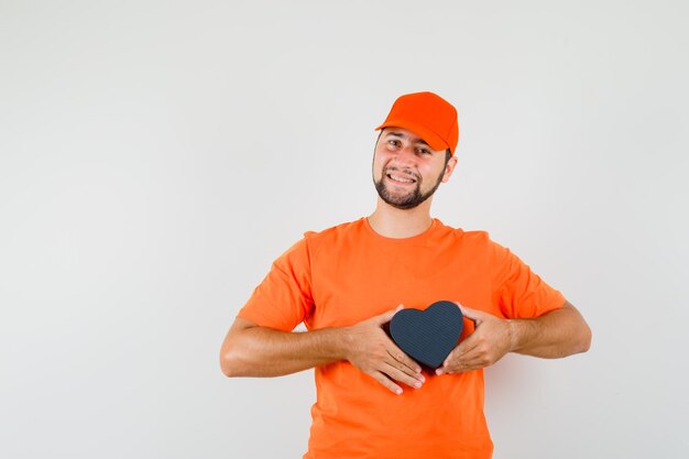 Delivery man holding gift box in orange t-shirt, cap and looking cheery. front view.