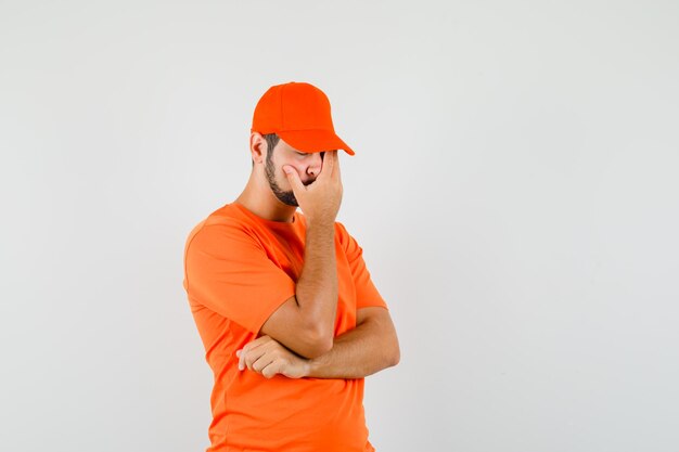 Delivery man holding hand on face in orange t-shirt, cap and looking regretful. front view.