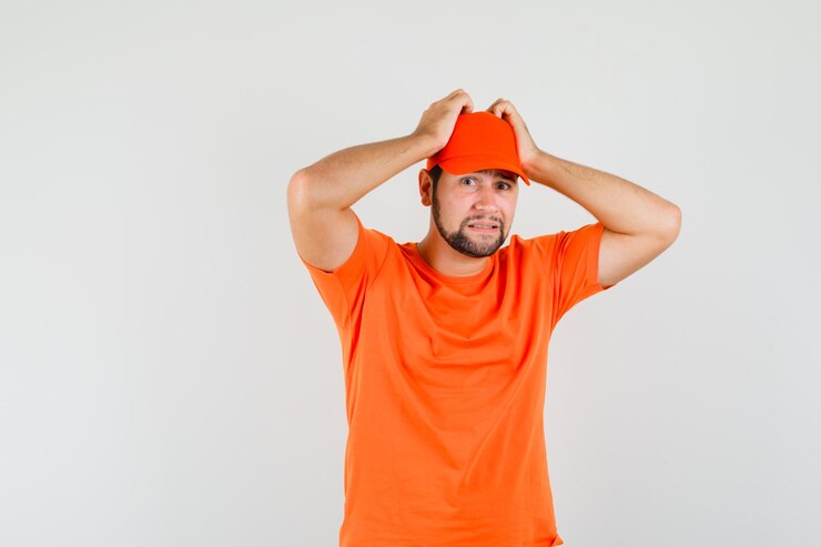 Free Photo delivery man holding hands on head in orange t-shirt, cap and looking puzzled , front view.
