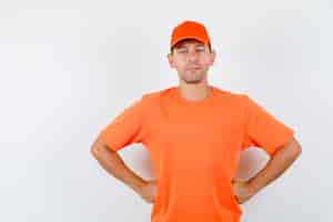 Free photo delivery man holding hands on waist and smiling in orange t-shirt and cap front view.