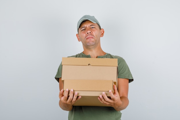 육군 녹색 t- 셔츠, 모자 전면보기에 무거운 골 판지 상자를 들고 배달 남자.