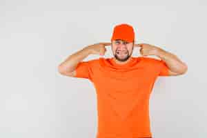 Бесплатное фото Курьер в оранжевой футболке, кепка затыкает уши пальцами и выглядит раздраженным, вид спереди.