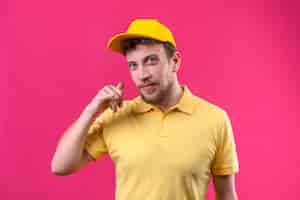 Бесплатное фото Курьер в желтой рубашке поло и кепке делает жест, который выглядит уверенно, дружелюбно улыбается на розовом
