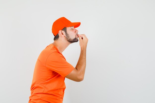 Курьер целует пальцы, делая восхитительный жест в оранжевой футболке, кепке и выглядит довольным. .