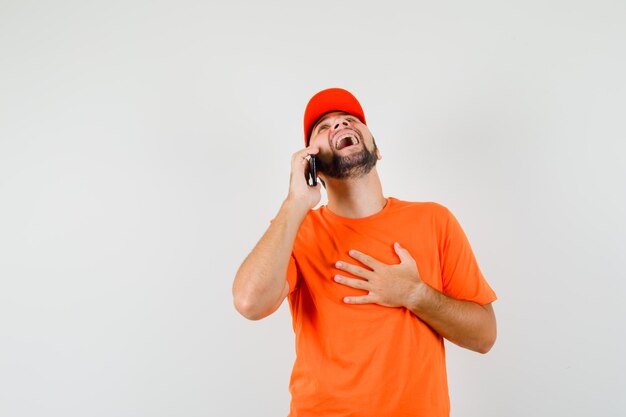 Delivery man laughing while talking on mobile phone in orange t-shirt, cap , front view.