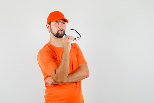 Delivery man looking up while holding glasses in orange t-shirt, cap and looking pensive , front view.