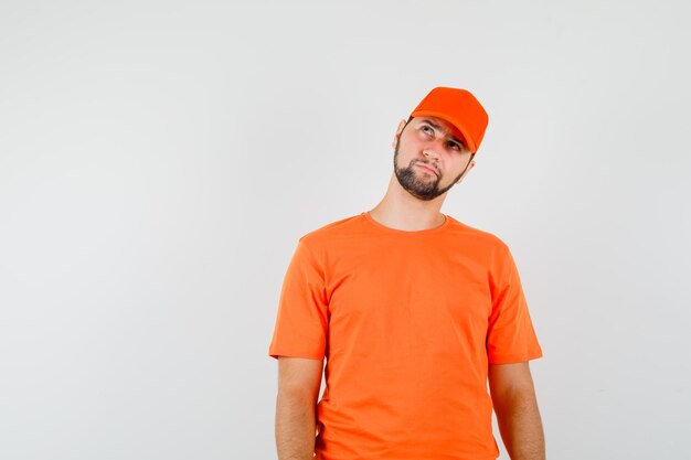 Delivery man looking upward in orange t-shirt, cap and looking pensive. front view.