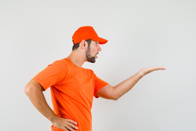 Доставщик делает вопросительный жест в оранжевой футболке, кепке и выглядит смущенным.