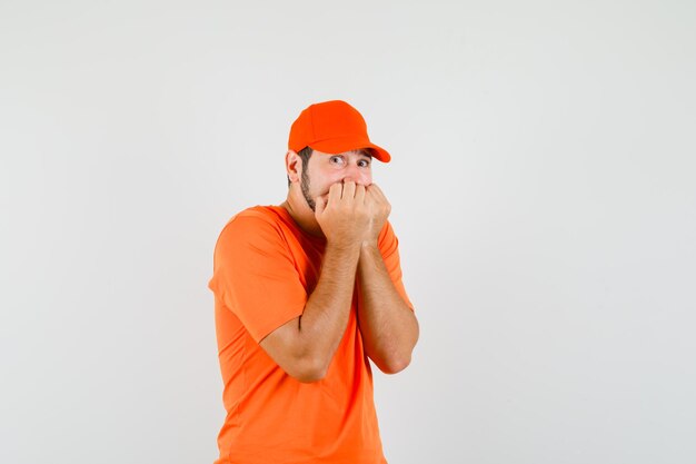 オレンジ色のTシャツを着た配達員、感情的に拳を噛み、怖がって見えるキャップ、正面図。