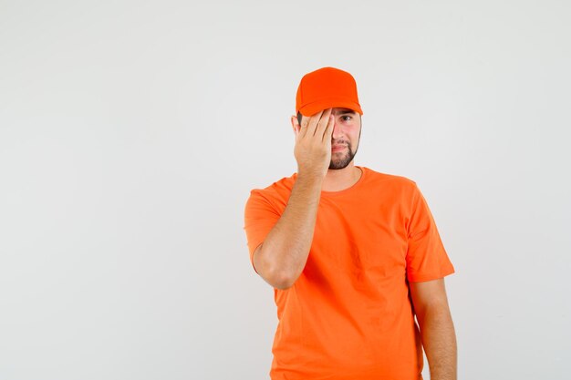 Доставщик в оранжевой футболке, крышка закрывает глаза рукой и выглядит позитивно, вид спереди.