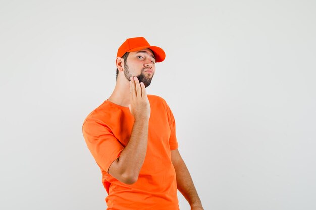 Delivery man in orange t-shirt, cap examining face skin by touching his beard , front view.