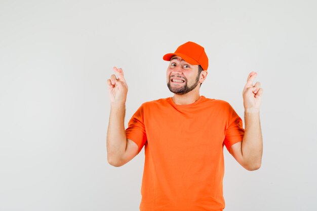 Delivery man in orange t-shirt, cap keeping fingers crossed and looking cheerful , front view.