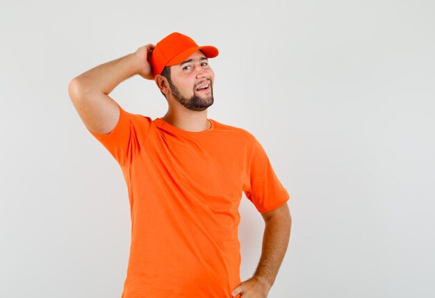 Delivery man in orange t-shirt, cap posing with hand behind head and looking elegant , front view.
