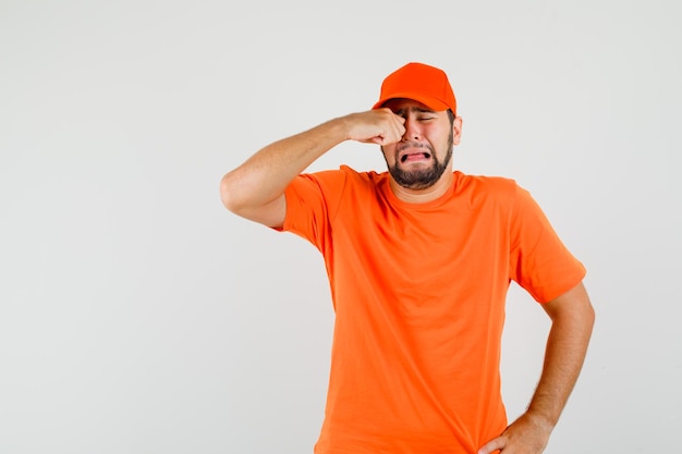 Delivery man in orange t-shirt, cap rubbing eye while crying like a child , front view.