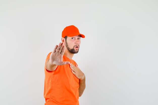 オレンジ色のTシャツを着た配達員、胸に手を当てて停止ジェスチャーを示し、怖がって見えるキャップ、正面図。